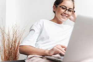 Image of smiling beautiful woman in eyeglasses using laptop while sitting on sofa at living room